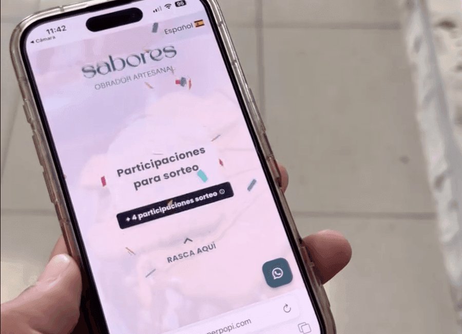 A close-up of a hand holding a smartphone displaying a mobile raffle interface for the brand 'sabores OBRADOR ARTESANAL' with confetti graphics, featuring the text 'Participaciones para sorteo' and a button labeled '+4 participaciones sorteo', along with a 'RASCA AQUÍ' prompt and a WhatsApp icon in the bottom right corner, set against a soft pink textured background with scattered confetti elements, while the phone’s status bar shows 11:42, 96% battery, and Spanish language setting.