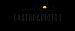 A minimalist logo design featuring a stylized frying pan with a yellow fried egg centered inside it, emitting small radiating lines to suggest heat or freshness, positioned above the word 'GASTROADICTOS' in bold, uppercase, sans-serif lettering with a slightly distressed or textured appearance, all rendered in white and yellow against a solid black background, conveying a modern, food-centric brand identity.