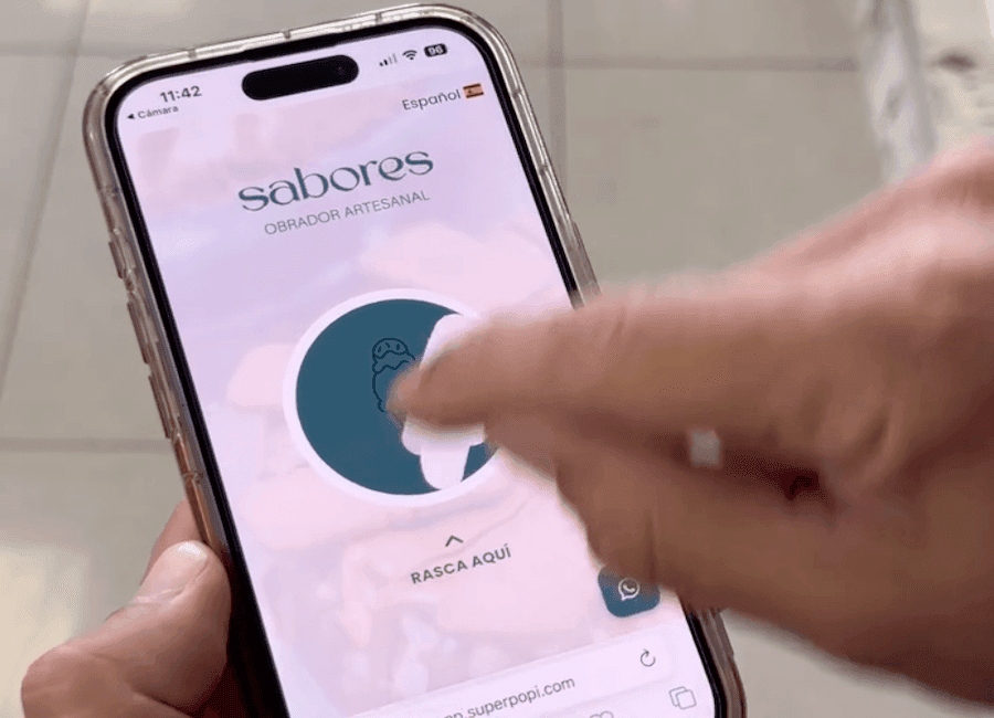 A close-up of a hand holding a smartphone with a clear protective case, displaying a mobile interface for the brand 'sabores OBRADOR ARTESANAL' in Spanish, featuring a circular teal scratch-off element with a small ice cream icon and the instruction 'RASCA AQUÍ' below it; a finger is actively scratching the surface, revealing part of the hidden content, while the phone’s status bar shows 11:42, 96% battery, and a Spanish language setting, with a WhatsApp icon and URL fragment 'superpopi.com' visible at the bottom.